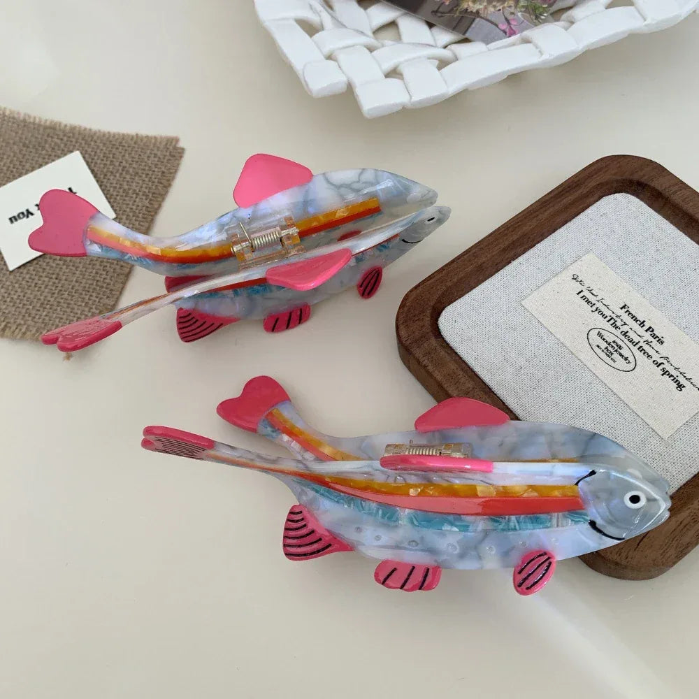 Two clear resin fish hair clips with pink fins and rainbow stripes arranged on a surface.