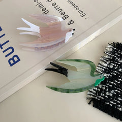 Two acrylic bird hair clips, one pearlescent pink and white, the other green and white, resting on a book with blue text.