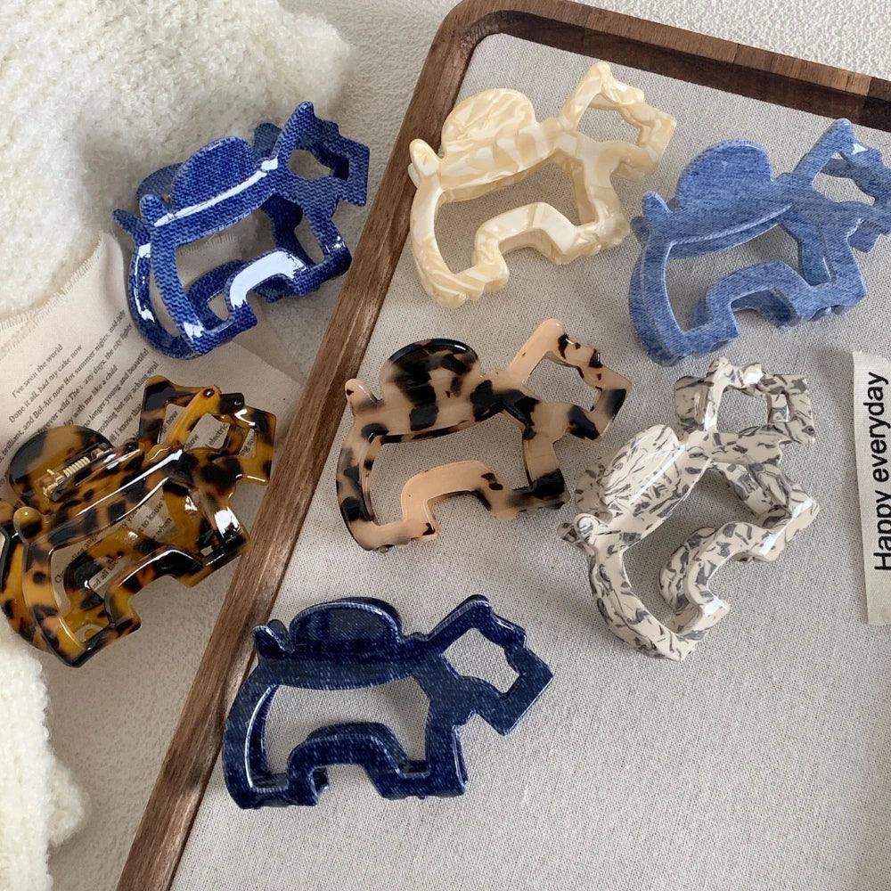 An assortment of elephant-shaped hair claw clips in various colors and patterns, laid out on a light-colored surface with a wooden tray.