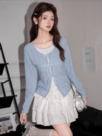 A young woman poses in a light blue cardigan and white ruffled skirt, holding a silver handbag.