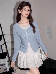 A young woman poses in a light blue cardigan and white ruffled skirt, holding a silver handbag.