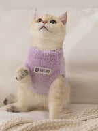 A light purple fuzzy cat sweater with a high neckline and a patch that says "WCAT" is worn by a cream-colored cat sitting down.