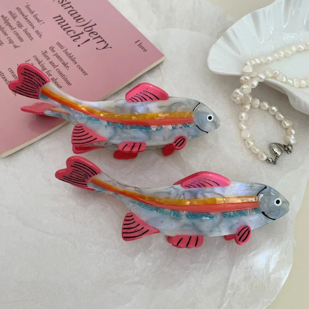 Two clear resin fish hair clips with pink fins and rainbow stripes beside a book and pearl necklace.