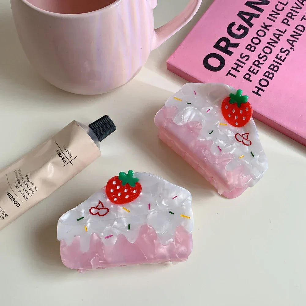 Two strawberry cake-shaped hair claw clips are displayed on a white surface next to a tube of hand cream and a pink book.