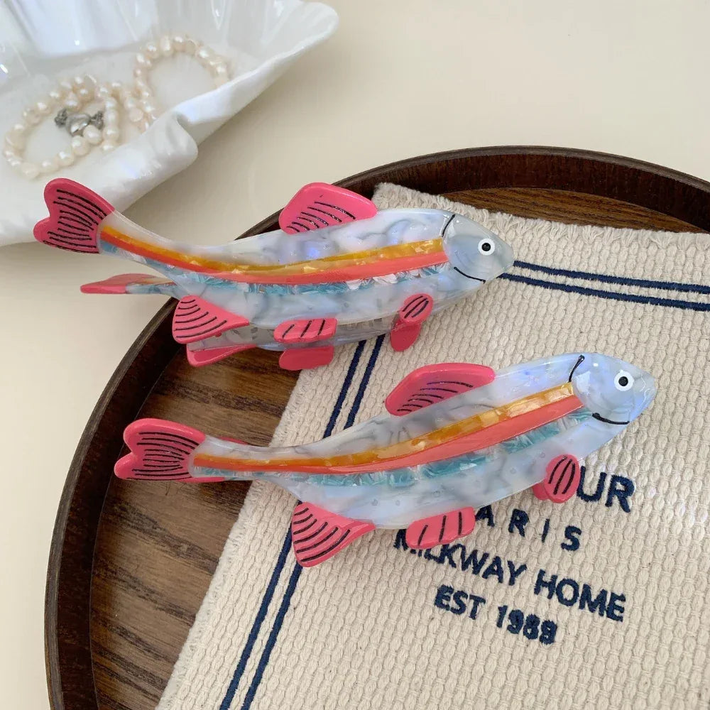 Two clear resin fish hair clips with pink fins and rainbow stripes placed on a wooden tray with a striped cloth.