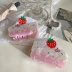 A pair of strawberry cake-shaped hair claw clips are placed on a white cloth with a pearl bracelet and a glass pedestal in the background.