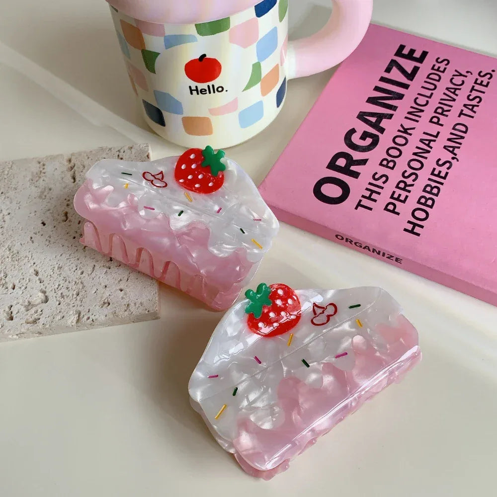 Two strawberry cake-shaped hair claw clips are arranged on a light-colored surface with a textured stone, a pink mug, and a pink book.