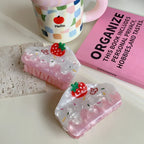 Two strawberry cake-shaped hair claw clips are arranged on a light-colored surface with a textured stone, a pink mug, and a pink book.