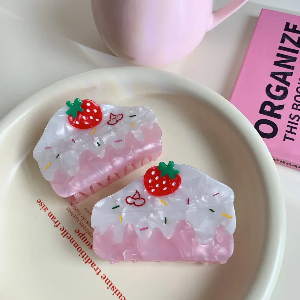 Two strawberry cake-shaped hair claw clips rest on a white plate with text, alongside a pink mug and a pink book in the background.