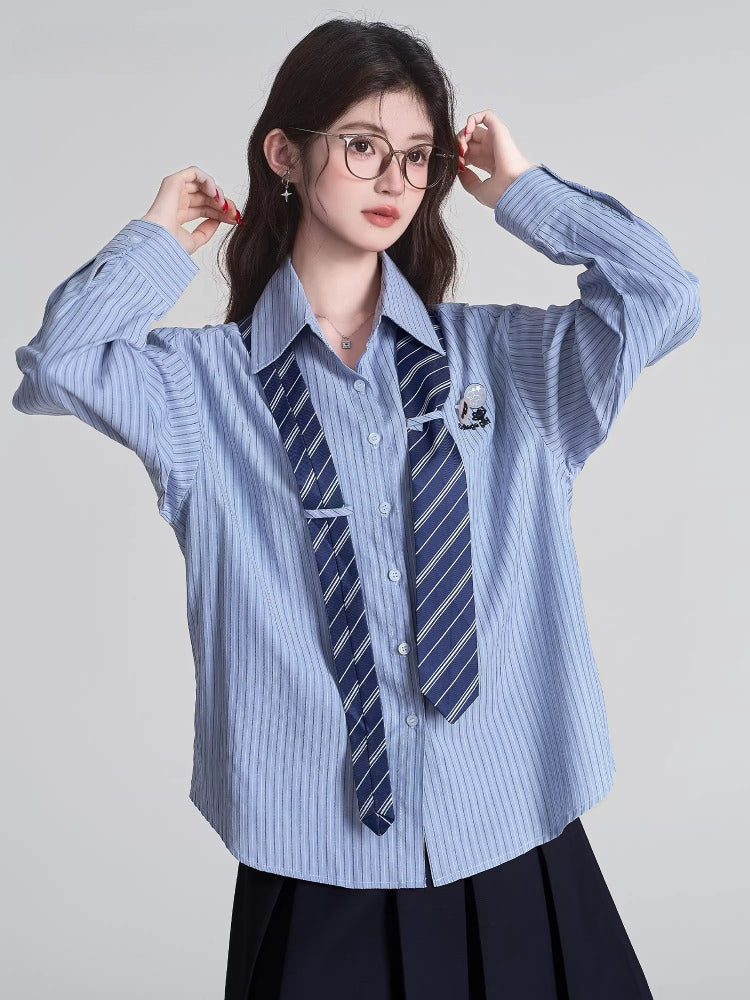 Model adjusting glasses while wearing a light blue striped button-down shirt with a navy striped tie and a black pleated skirt.