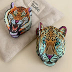 A tiger head accessory and a leopard head accessory displayed on a textured fabric background