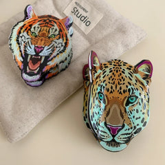 A tiger head accessory and a leopard head accessory displayed on a textured fabric background