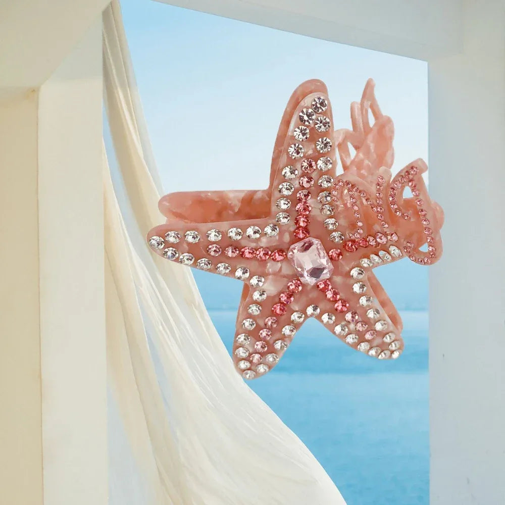 A pink rhinestone-embellished starfish hair claw clip against a backdrop of sheer white fabric and a blue ocean scene