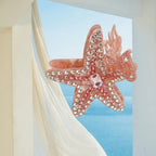 A pink rhinestone-embellished starfish hair claw clip against a backdrop of sheer white fabric and a blue ocean scene