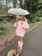 Woman with blonde hair in a ponytail, wearing a pink chunky knit sweater, white lace shorts, and pink rain boots, holding a mug and an umbrella.