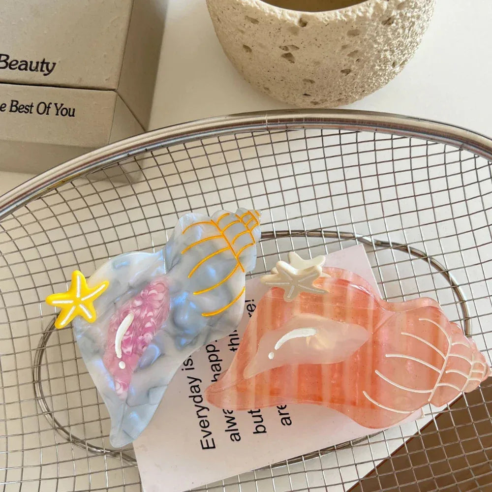 Two seashell-shaped hair clips, one pink and one blue/grey, with starfish accents, resting on a piece of paper inside a wire basket.
