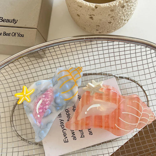 Two seashell-shaped hair clips, one pink and one blue/grey, with starfish accents, resting on a piece of paper inside a wire basket.