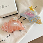 Two seashell-shaped hair clips, one pink and one blue/grey, with starfish accents, placed on a beige cloth.
