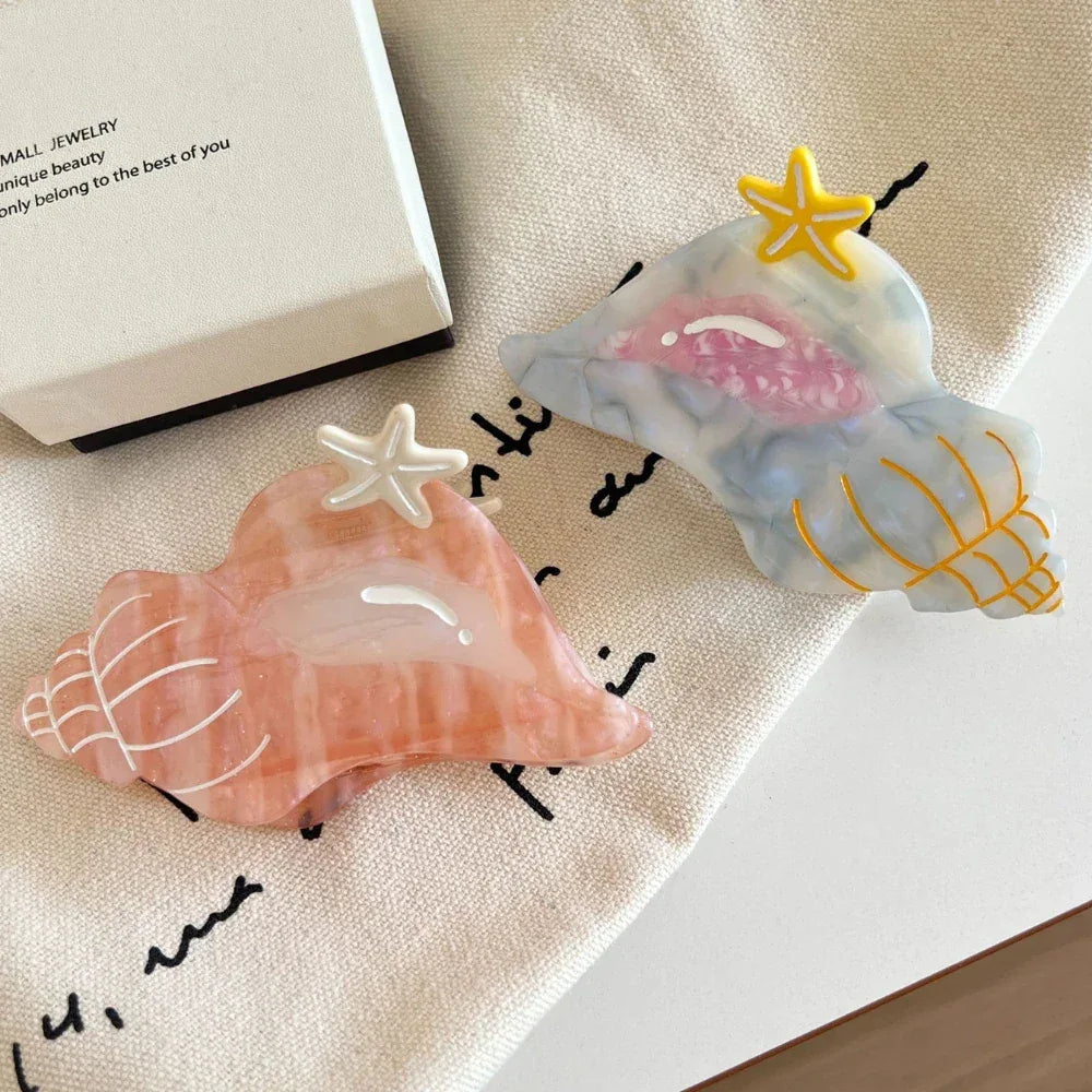 Two seashell-shaped hair clips, one pink and one blue/grey, with starfish accents, placed on a beige cloth.