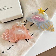 Two seashell-shaped hair clips, one pink and one blue/grey, with starfish accents, placed on a beige cloth.