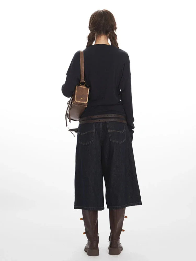 Back view of a woman wearing a dark long-sleeved V-neck top, dark wide-leg cropped denim pants, a brown leather belt, brown boots, and a brown leather crossbody bag, posing against a white background.