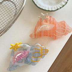 Two seashell-shaped hair clips, one pink and one blue/grey, with starfish accents, arranged on a white surface.