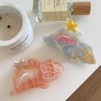 Two seashell-shaped hair clips, one pink and one blue/grey, with starfish accents, placed near a candle and a bottle of perfume on a white surface.