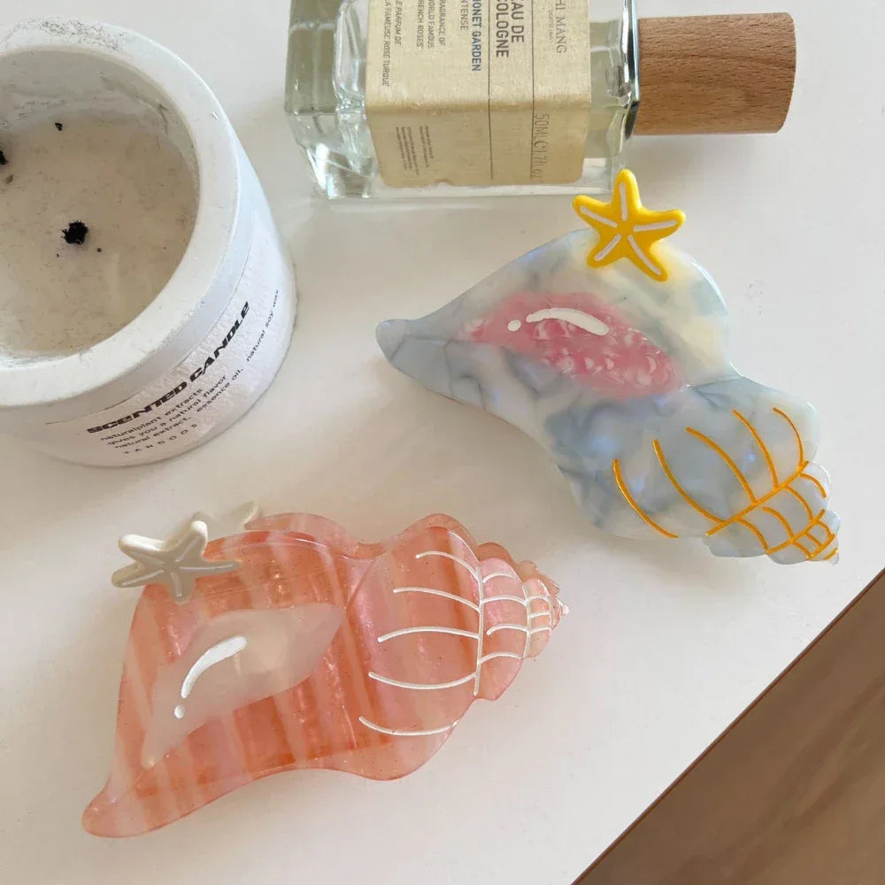 Two seashell-shaped hair clips, one pink and one blue/grey, with starfish accents, placed near a candle and a bottle of perfume on a white surface.