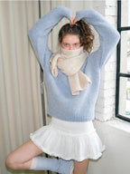 Model in a light blue knit sweater, cream scarf, and white ruffled skirt, posing with arms raised.
