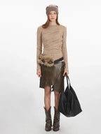 Woman wearing a beige long-sleeve knit top, an olive green faux leather skirt with an asymmetrical hem, a beige beanie, and brown boots, holding a black tote bag.