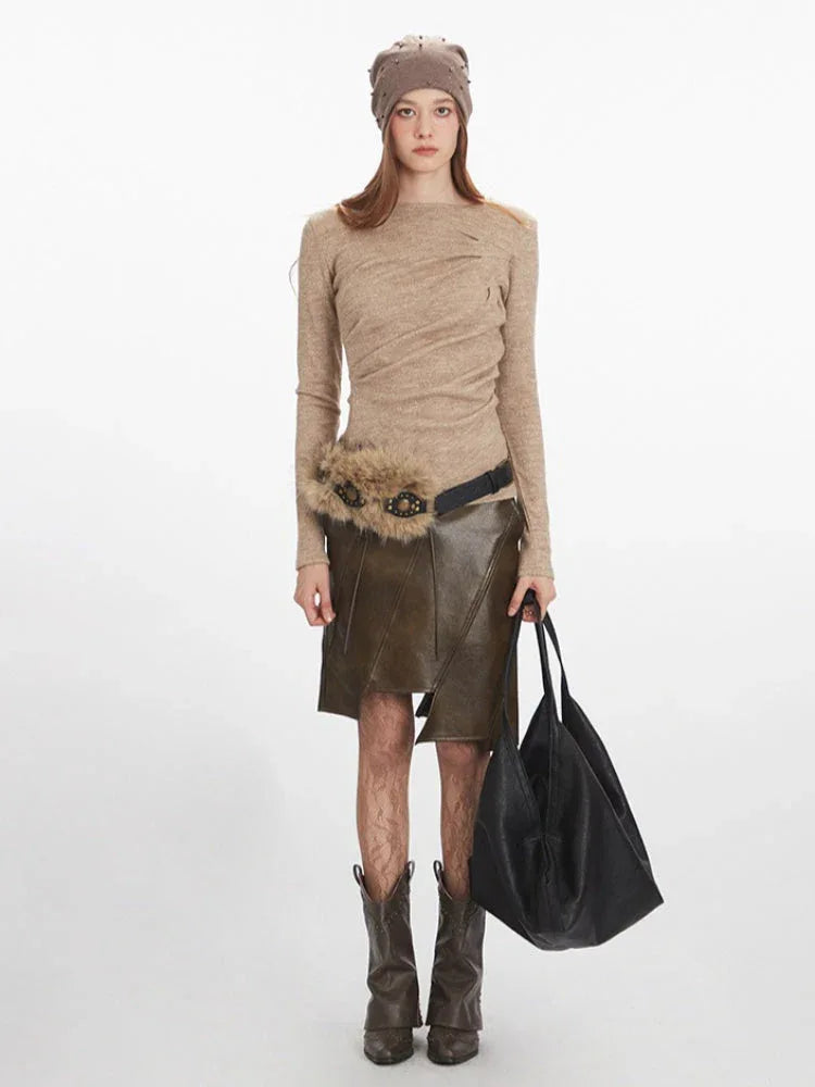 Woman wearing a beige long-sleeve knit top, an olive green faux leather skirt with an asymmetrical hem, a beige beanie, and brown boots, holding a black tote bag.