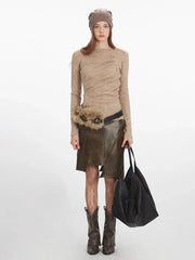 Woman wearing a beige long-sleeve knit top, an olive green faux leather skirt with an asymmetrical hem, a beige beanie, and brown boots, holding a black tote bag.