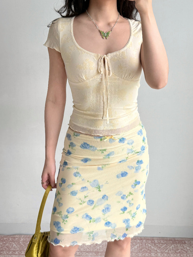 Full body shot of a woman wearing a pale yellow floral print mesh top and a matching skirt, holding a green handbag.