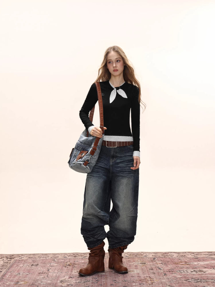 Full body shot of a model wearing a black long-sleeve top with a white polka dot scarf detail and white polka dot cuffs, paired with baggy denim jeans, a brown belt, brown boots, and a denim crossbody bag.