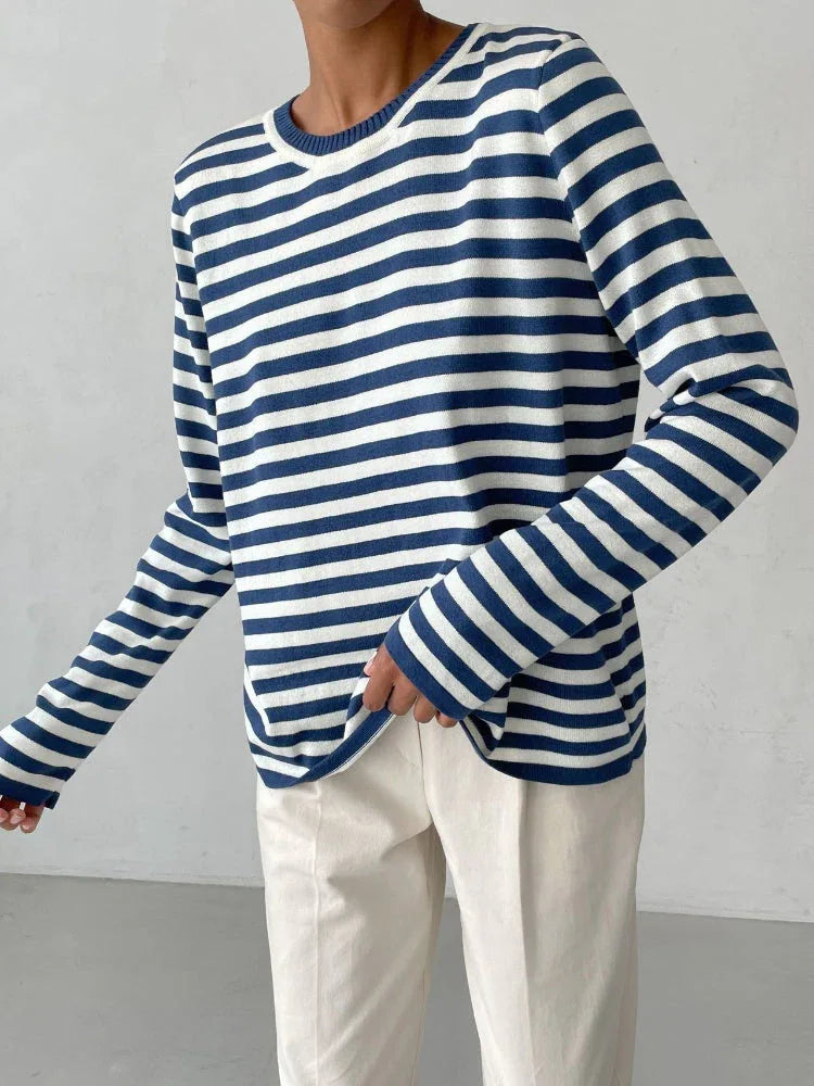 Close-up of a woman wearing a blue and white striped long-sleeved crew neck sweater with cream-colored pants, holding the hem of the sweater.