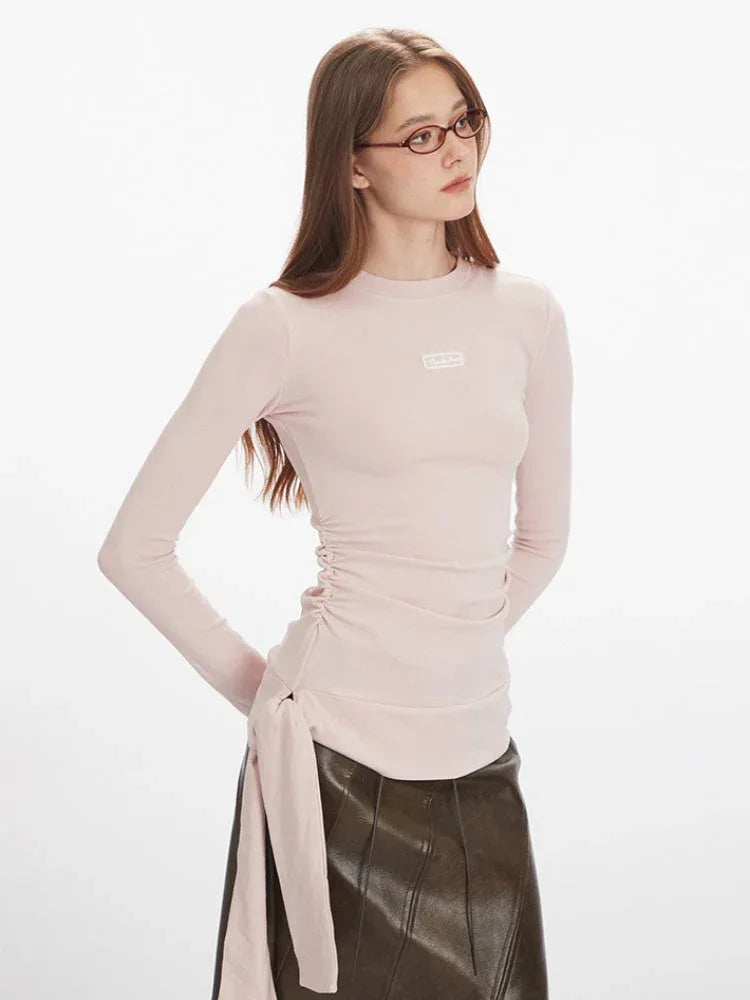 Close-up front view of a woman wearing a pink long-sleeve ruched top with a tie detail and a brown asymmetrical faux leather skirt.