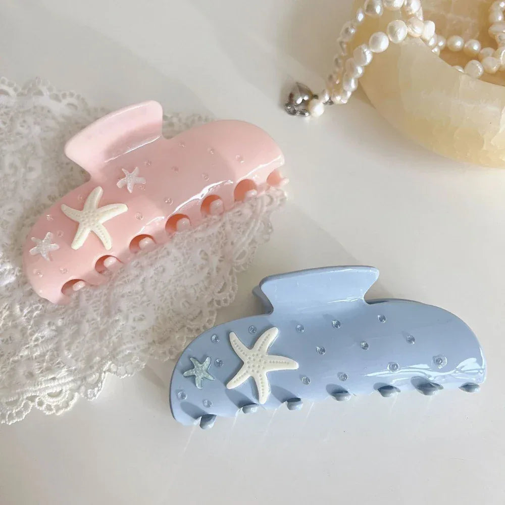 two pastel claw clips, one pink and one light blue, both with starfish and rhinestones, on a white lace tablecloth