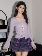 Close-up of a woman wearing a lavender ribbed knit cardigan with lace trim and bow details over a white camisole, paired with a purple tiered ruffled skirt.