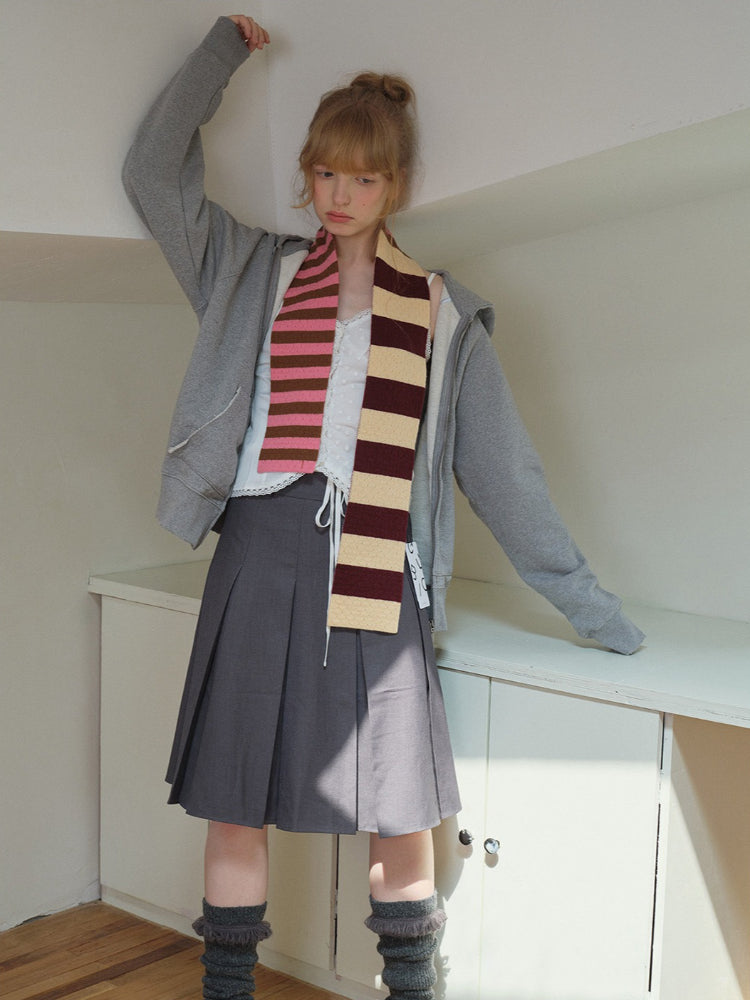 Model posing with arms raised, wearing a grey zip-up hoodie, grey pleated skirt, and a striped scarf.