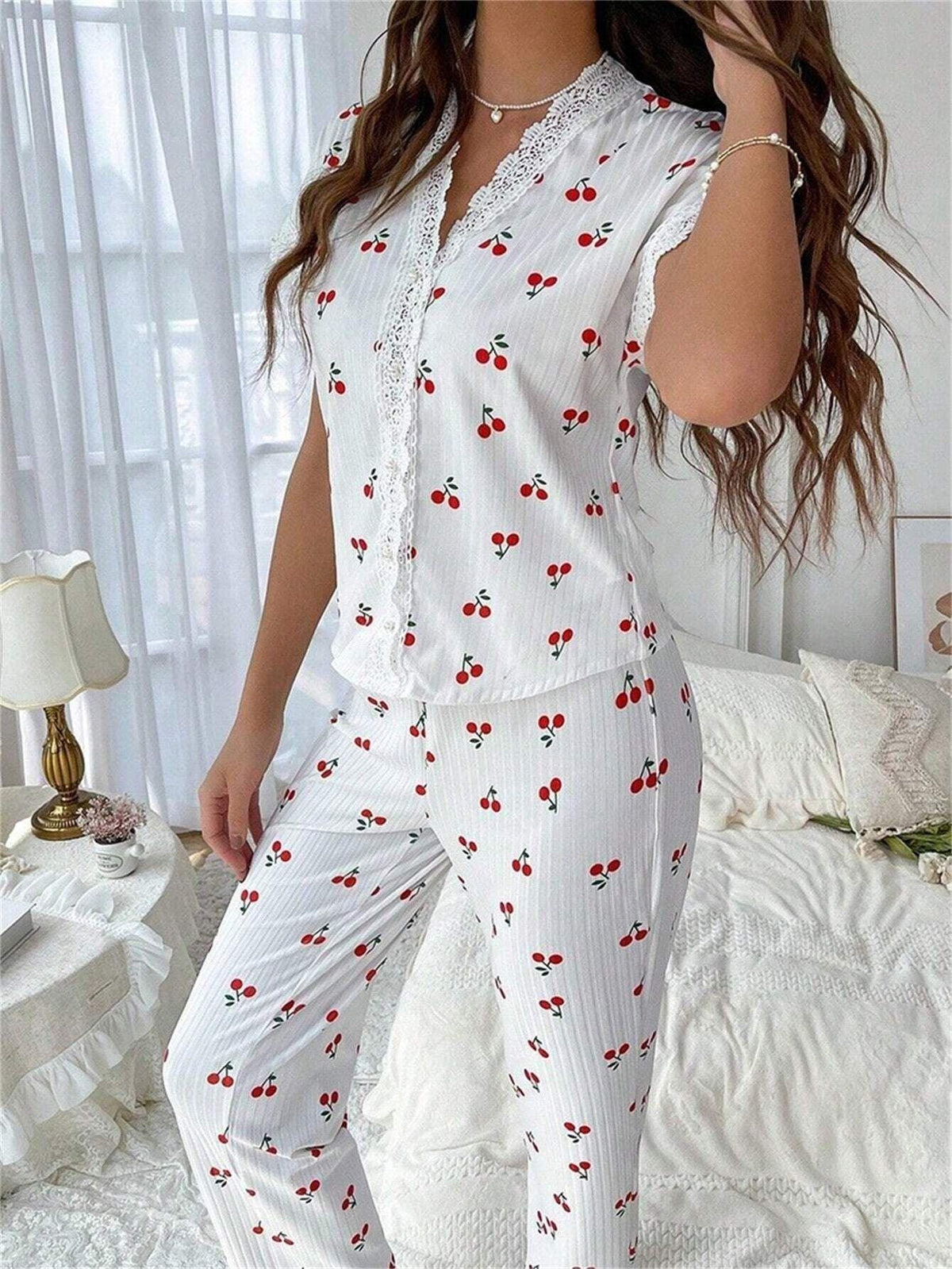 Close-up of a woman wearing a white short-sleeve pajama top with cherry print and white ribbed pants with cherry print, standing in a bedroom.