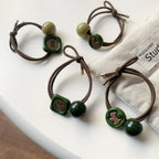 Four hair ties with decorative charms and beads, shown with a white fabric item in the background.