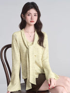 Woman sitting on a chair, wearing a light yellow textured knit top with a draped neckline and scarf tie.