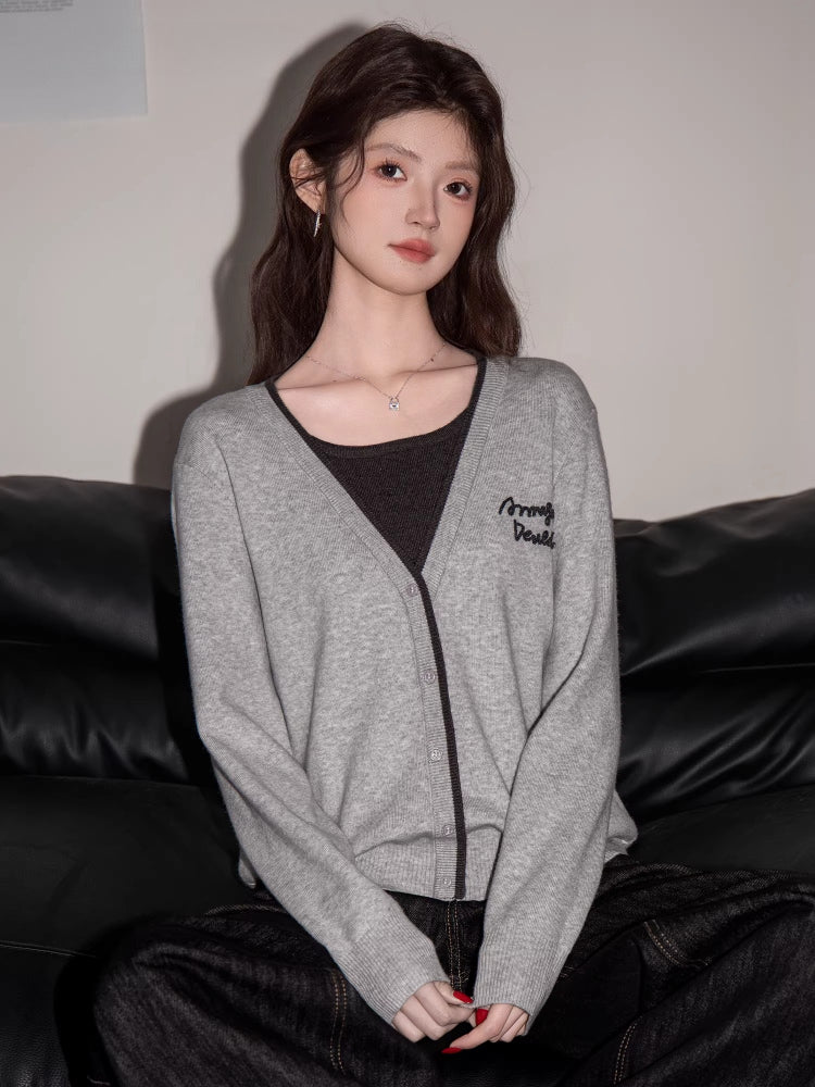 Front view of a woman in a grey knit cardigan with black trim and embroidery, dark top, and dark wide-leg jeans, seated on a black couch.