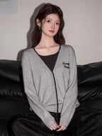 Front view of a woman in a grey knit cardigan with black trim and embroidery, dark top, and dark wide-leg jeans, seated on a black couch.