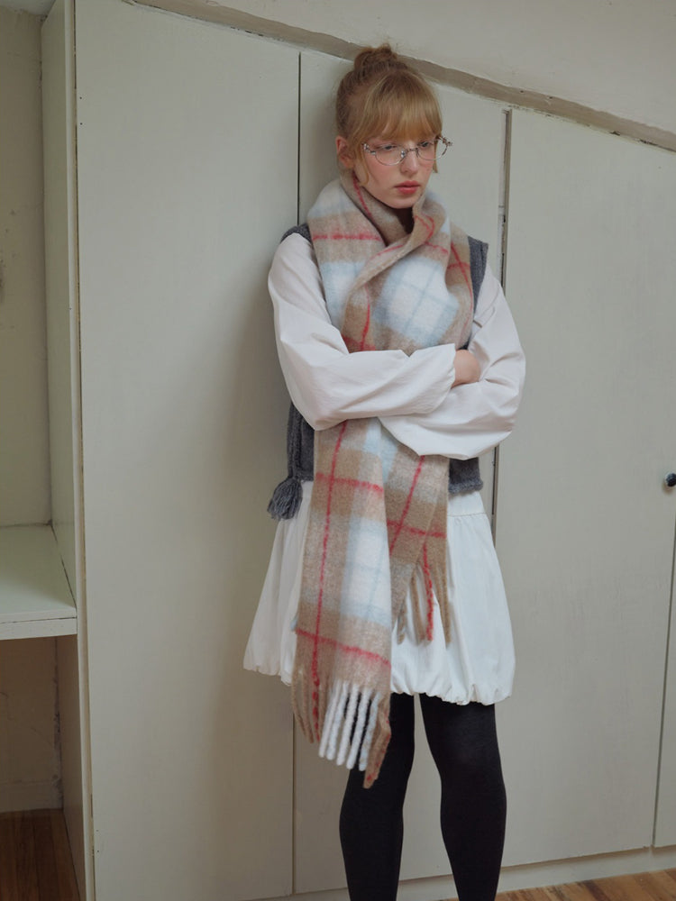 Woman standing with arms crossed, wearing a plaid scarf over a white dress and grey vest.