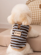 Back view of a white bichon frise dog wearing a blue and brown striped fleece vest with sherpa trim and a logo patch