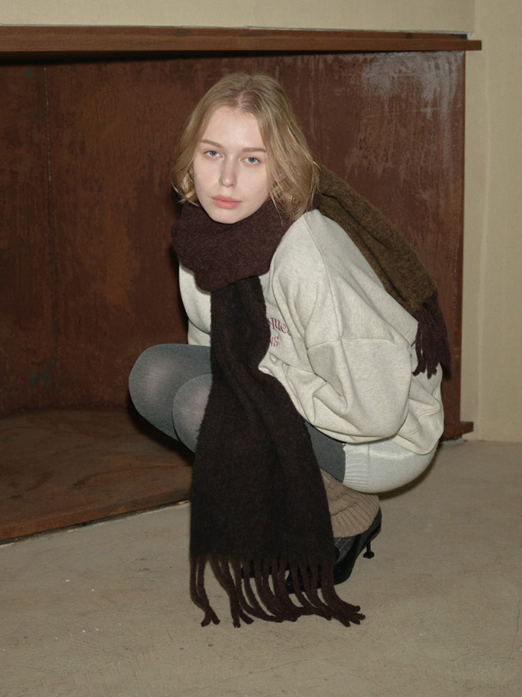 A young person wearing a cream-colored crew neck sweatshirt and a dark brown fringed scarf, crouching down.