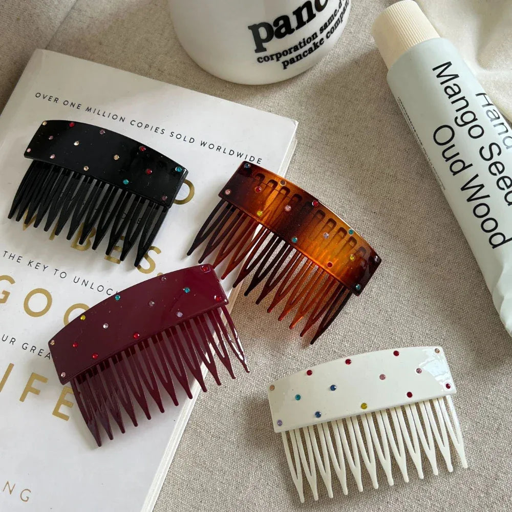 Four hair combs in different colors (black, tortoiseshell, burgundy, and white) arranged on a book and fabric.