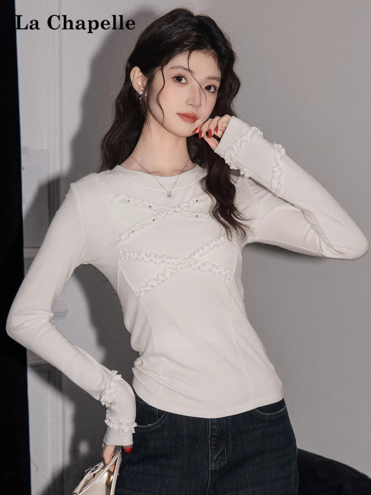 Woman posing in a white long-sleeved top featuring ruffled neckline details and beaded accents, with dark wash jeans.