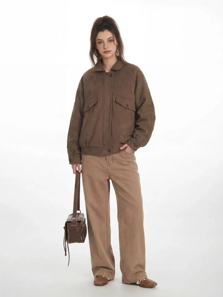 Woman wearing a brown bomber jacket and tan pants, with one hand in her pocket, holding a brown bag.